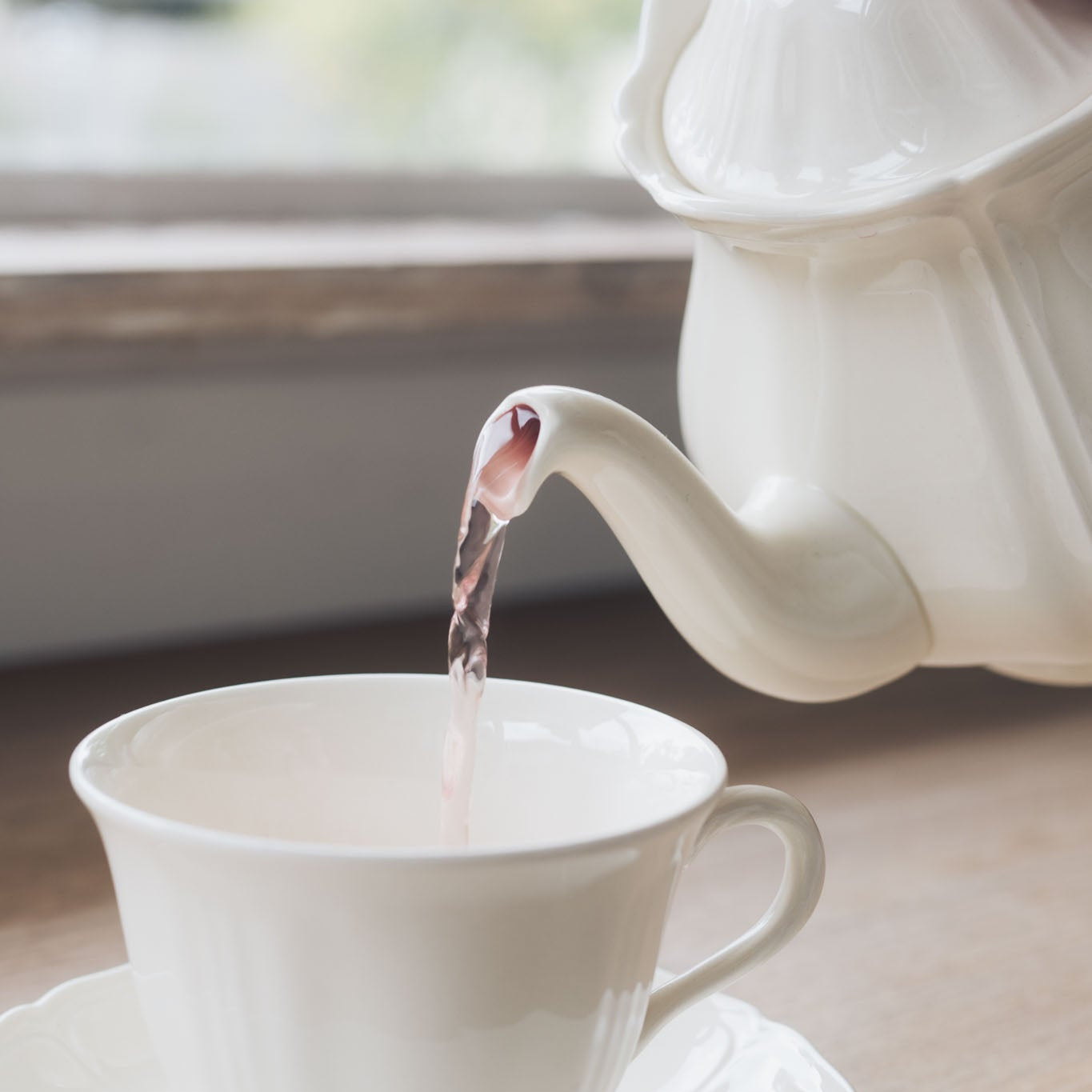 White teapot pouring tea into a white teacup on a wooden table.
