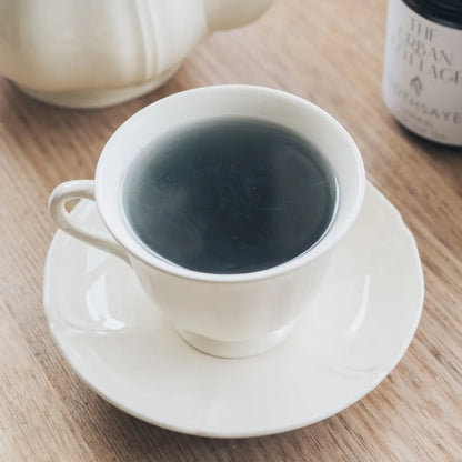 A vintage cream coloured cup and saucer with tea filled to the brim of a blue hue, matching teapot and glass jar of Soothsayer tea in background