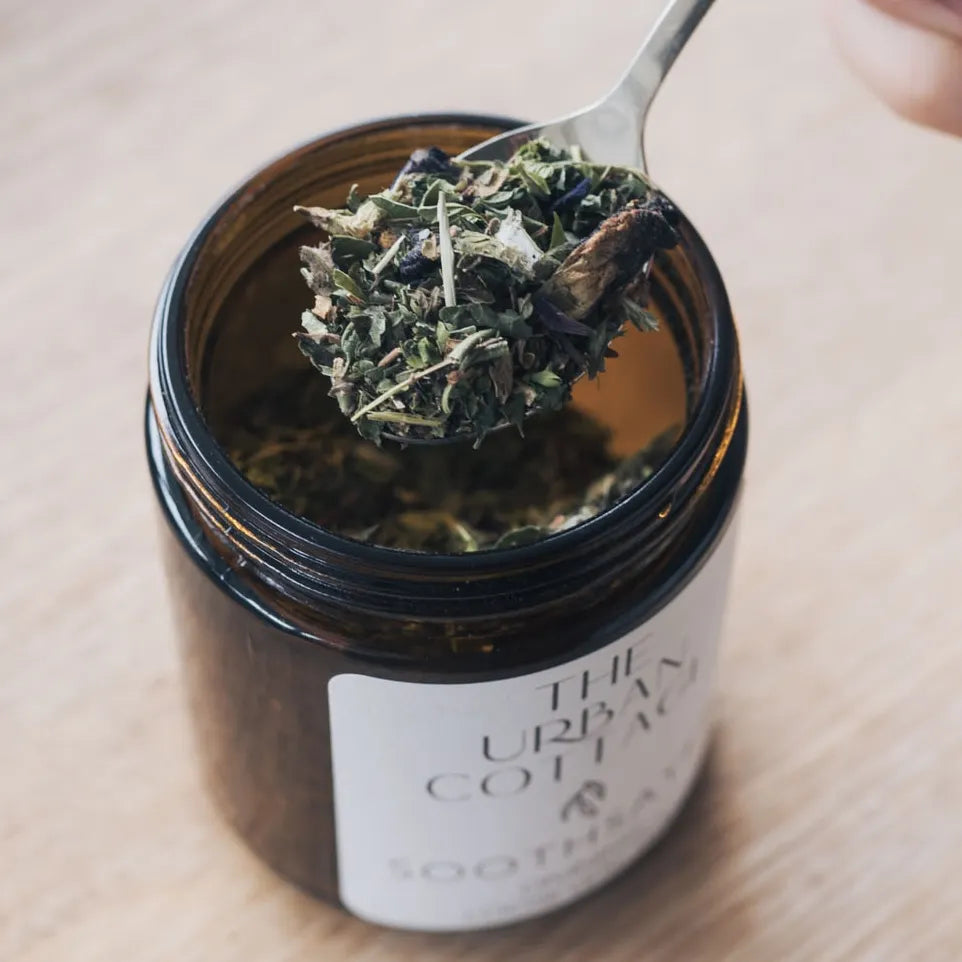 A scoop of loose tea leaves and flowers heaped on a teaspoon above an open amber jar of Soothsayer Divination Blend tea.