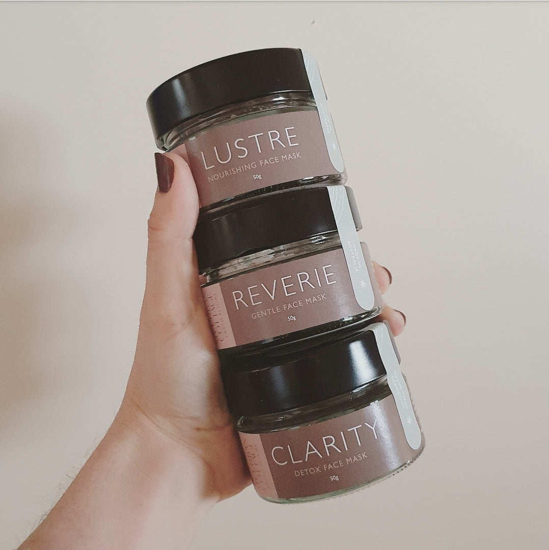 Female hand with painted nail holding 3 stacked jars of face masks labelled "LUSTRE", "REVERIE" and "CLARITY".