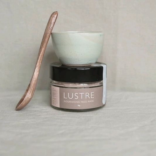 Screw top glass jar with label reading "LUSTRE Nourishing Face Mask" with small bowl on top and wooden spoon learning against it.